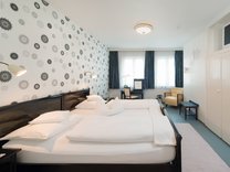 Modern eingerichtetes Hotelzimmer mit zwei nebeneinander stehenden Betten, großem Fenster mit Vorhängen und einer Wand mit dekorativem Muster