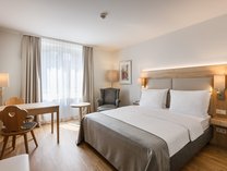 Hotelzimmer mit großem Doppelbett, zwei Nachttischen, Schreibtisch mit Stuhl und Fenster mit Vorhängen
