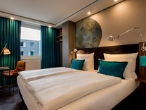 Modern eingerichtetes Hotelzimmer mit Doppelbett, zwei türkisfarbenen Kissen, Nachttischlampen und Fenster mit grünen Vorhängen