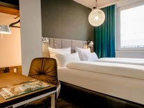 Hotelzimmer mit großem Bett, Schreibtisch mit braunem Stuhl und runder Deckenlampe