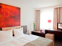 Hotelzimmer mit Doppelbett, großem roten Gemälde über dem Bett, Schreibtisch mit Stuhl und Fernseher vor einem Fenster mit Vorhängen