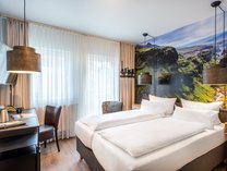 Hotelzimmer mit zwei Einzelbetten, Schreibtisch mit Stuhl, großen Fenstern mit Vorhängen und einer Wand mit Landschaftsfotografie als Tapete